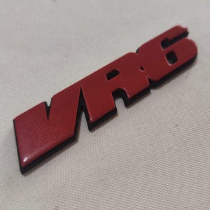 Könnte beinhalten: Ein rot-schwarzes "VR6" Emblem. Das Emblem hat eine glänzende rote Oberfläche mit schwarzer Kontur. Die Buchstaben sind stilisiert und scheinen aus Metall oder einem ähnlichen Material zu bestehen. Das Emblem ist wahrscheinlich für ein Fahrzeug.