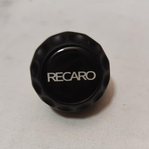 Puede incluir: Pomo redondo negro con una superficie texturizada y la palabra "RECARO" impresa en blanco.