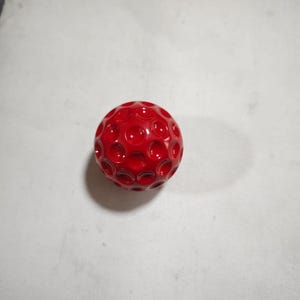 Könnte beinhalten: Eine glänzende, rote Kugel mit einer genoppten Oberfläche, die an einen Golfball erinnert. Das Objekt ist ein sattes, leuchtendes Rot, und die Vertiefungen erzeugen Schatten, die Tiefe verleihen. Die Kugel befindet sich vor einem schlichten, cremefarbenen Hintergrund.