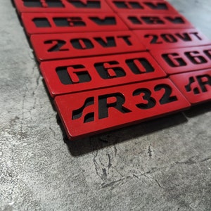 May include: A stack of red metal plates with black lettering. The plates have different text on them, including "GT", "VW", "20VT", "G60", and "R32".