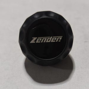 May include: Black round knob with a textured surface and the word "Zender" printed in white.
