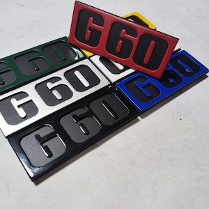 Peut inclure: Plusieurs emblèmes rectangulaires avec le numéro "G60" en différentes couleurs, dont le rouge, le vert, l'argent, le noir et le bleu. Le texte "G60" est en relief et dans une couleur contrastante avec le fond.