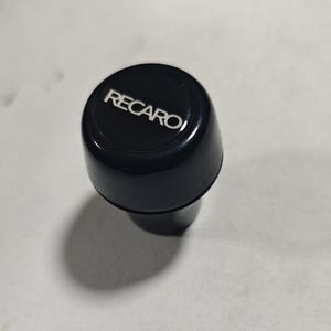 Może przedstawiać: Czarny gałka zmiany biegów z białym napisem "RECARO" na górze. Gałka ma zaokrągloną górę i zwężającą się podstawę. Zdjęcie zrobione jest z lekko podwyższonego kąta.