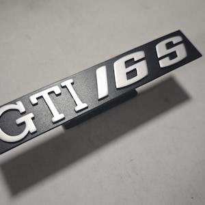 Może przedstawiać: Czarny, prostokątny emblemat z napisem "GTI 16S" w srebrnych literach. Litery są wypukłe i wyróżniają się na ciemnym tle. Emblemat jest lekko pochylony, rzucając cień.