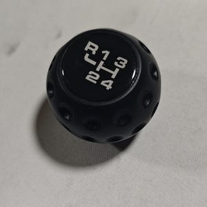 Könnte beinhalten: Schwarzer Schaltknauf mit weißen Buchstaben, die die Gangpositionen anzeigen: R, 1, 2, 3 und 4. Der Knauf hat eine runde Form mit kleinen kreisförmigen Vertiefungen.