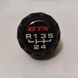 May include: Black gear shift knob with a white and red 'GTX' logo and white numbers 'R135LHL24' on the top.