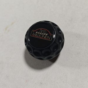 May include: A black, spherical gear shift knob with a dimpled, golf ball-like texture. The top center features a flat, circular emblem with the text 19 GOLF MILLIONEN in white and red lettering against a black background.