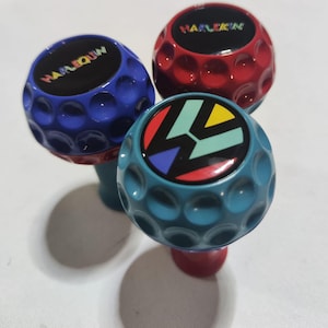Pu&ograve; includere: Tre pomelli a forma di palla da golf con colori e design diversi. Il primo pomello &egrave; blu con la parola "HARLEQUIN" in lettere arcobaleno. Il secondo pomello &egrave; rosso con la parola "HARLEQUIN" in lettere bianche. Il terzo pomello &egrave; blu-verde con un design geometrico colorato.