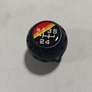Może przedstawiać: Czarny gałka zmiany biegów z czerwonym, żółtym i czarnym akcentem. Wzór zmiany biegów jest wyświetlany białym tekstem: R, 1, 3, 5, H, 2, 4. Gałka ma teksturowaną powierzchnię.