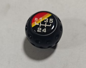 May include: Black gear shift knob with a red, yellow, and black accent. The shift pattern is displayed in white text: R, 1, 3, 5, H, 2, 4. The knob has a dimpled texture.