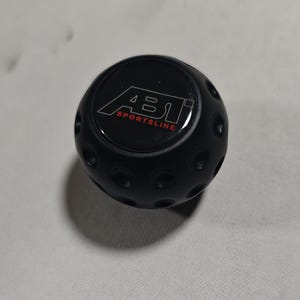 May include: Black shift knob with the text "ABT SPORTSLINE" in red and white. The knob has a rounded shape with small circular indentations around the sides. The product is likely an automotive accessory.