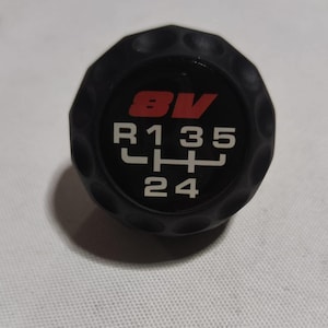 以下が含まれることがあります： 白い数字と赤い8Vのロゴが付いた黒いシフトノブ。ノブの数字はR、1、3、5、L、H、2、4です。