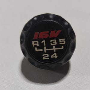 以下が含まれることがあります： 白と赤の文字が書かれた黒いシフトノブ。文字は「16V」と「R1 3 5 L H 2 4」です。