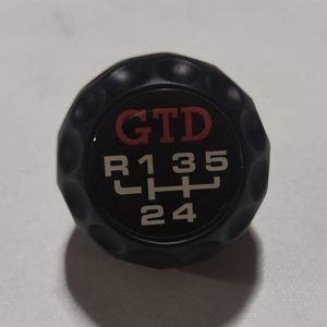 Può includere: Pomello del cambio nero con logo "GTD" rosso e numeri bianchi "R135 24" stampati su di esso.