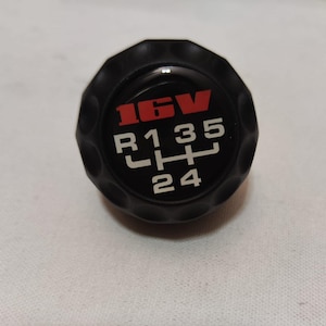 Puede incluir: Pomo de cambio de marchas negro con un logotipo 16V blanco y rojo y números blancos 1, 3, 5, R, 2 y 4.