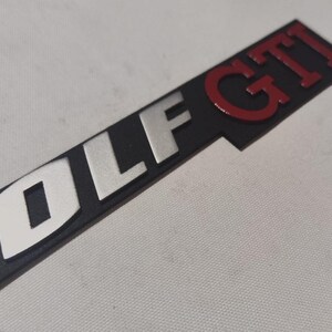 Könnte beinhalten: Ein schwarzes Golf GTI-Emblem mit silbernen und roten Buchstaben. Das Wort "GOLF" ist silberfarben und "GTI" ist rot. Das Emblem ist rechteckig und hat eine strukturierte Oberfläche.