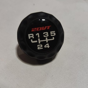 Könnte beinhalten: Schwarzer Schaltknauf mit weißen Zahlen und einem roten "20VT"-Logo. Die Zahlen auf dem Knauf sind R, 1, 3, 5, 2 und 4.