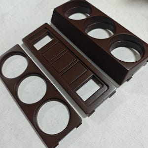 Può includere: Tre componenti in plastica marrone scuro per il cruscotto di un veicolo. Un pezzo presenta tre ritagli circolari, un altro è una cornice rettangolare con quattro slot per pulsanti e il terzo è un alloggiamento rialzato con tre aperture.