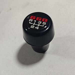 Könnte beinhalten: Schwarzer Schaltknauf mit einem roten "G60" und weißen Zahlen und Buchstaben, die die Gangpositionen angeben: "R135 24".