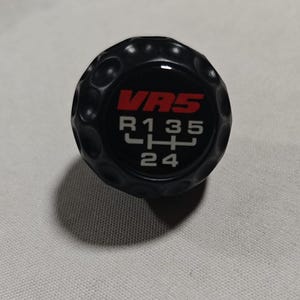 May include: A black spherical gear shift knob with a dimpled texture. The top surface features the red text VR5 above a white gear shift pattern showing R, 1, 3, 5, 2, and 4 positions. The knob has a smooth, glossy center inlay surrounded by a textured grip.