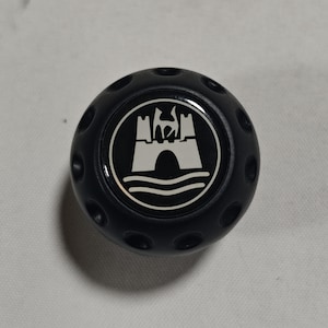 Puede incluir: Un pomo negro y redondo con un emblema circular que presenta un diseño de castillo blanco. El pomo tiene varios agujeros pequeños y espaciados uniformemente alrededor de su perímetro. El diseño del castillo incluye un puente y líneas onduladas.