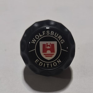 Peut inclure: Emblème rond noir avec le texte "Wolfsburg Edition" en lettres blanches autour d'un blason noir et blanc avec un château rouge à l'intérieur.