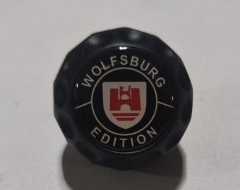 Puede incluir: Emblema redondo negro con el texto "Wolfsburg Edition" en letras blancas alrededor de un escudo negro y blanco con un castillo rojo en su interior.