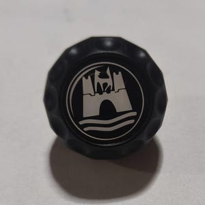 Könnte beinhalten: Schwarzes Plastik-Autoemblem mit einem silbernen Volkswagen-Logo mit einem Schloss und Wellen.