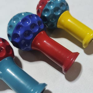 Pu&ograve; includere: Tre giocattoli a forma di palla da golf con basi di colori diversi. Le palle da golf sono rosse e blu, rosse e verde acqua, e verde acqua e gialle. Le basi sono rosse, verde acqua e gialle.