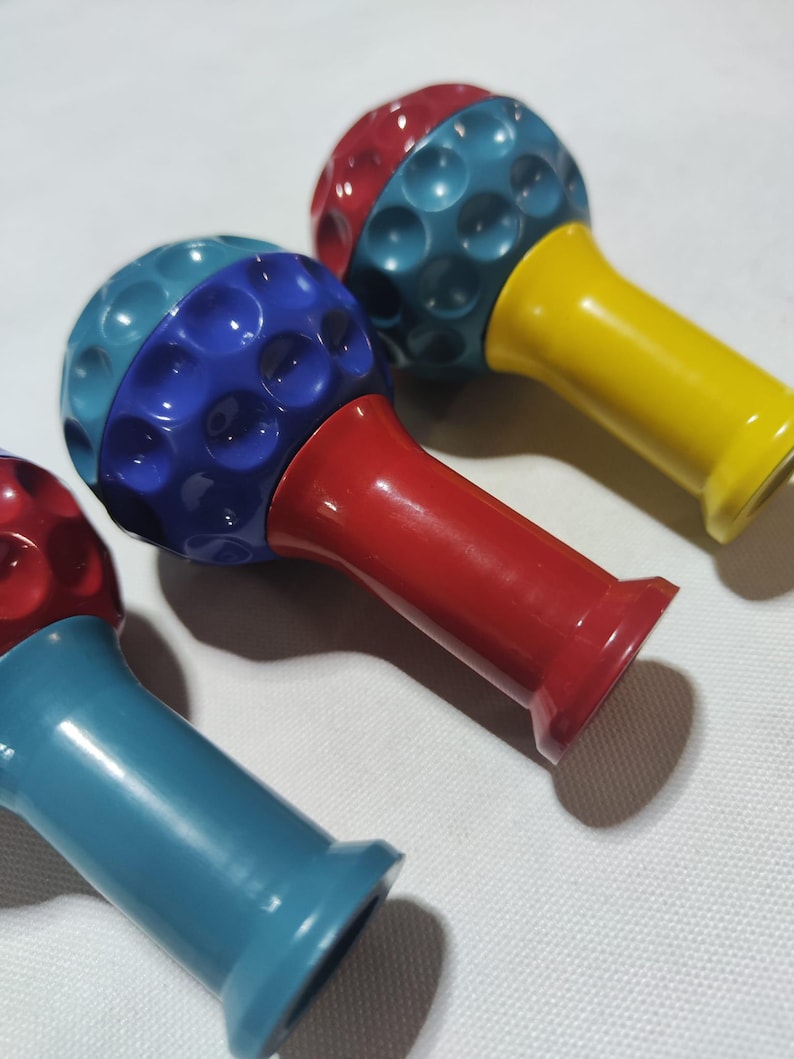 Pu&ograve; includere: Tre giocattoli a forma di palla da golf con basi di colori diversi. Le palle da golf sono rosse e blu, rosse e verde acqua, e verde acqua e gialle. Le basi sono rosse, verde acqua e gialle.