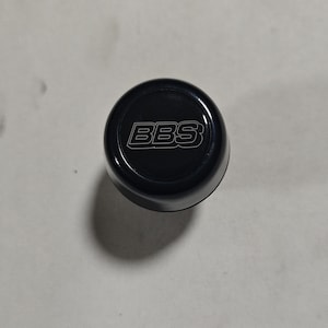 Peut inclure: Un bouchon de valve rond noir avec le mot "BBS" imprimé en blanc.