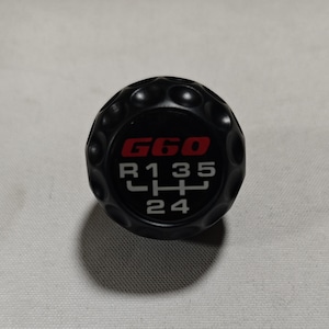 Può includere: Pomello del cambio nero con la scritta "G60" in rosso e un motivo del cambio bianco. Il pomello ha una superficie strutturata ed è progettato per un cambio manuale.