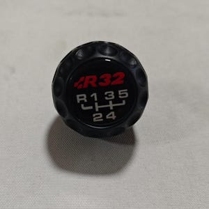 Puede incluir: Pomo de palanca de cambios negro con el texto "R32" en rojo y blanco, y un diagrama de patrón de marchas. El pomo tiene una superficie texturizada y está diseñado para la transmisión manual de un vehículo.