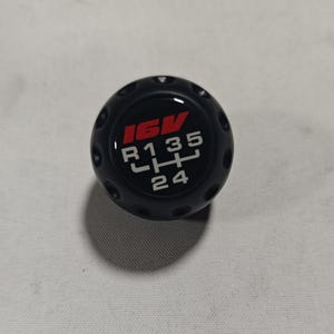 May include: Black gear shift knob with a red and white graphic. The graphic displays the text "16V" in red, and "R 1 3 5" and "2 4" in white, indicating the gear positions. The knob has a textured surface with small circular indentations.