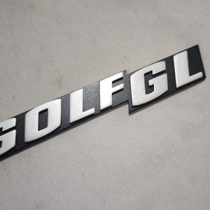 Könnte beinhalten: Ein schwarz-silbernes "GOLF GL" Emblem. Die Buchstaben sind silberfarben und erhaben, vor einem schwarzen rechteckigen Hintergrund. Das Emblem ist leicht angewinkelt, mit strukturierter Oberfläche.