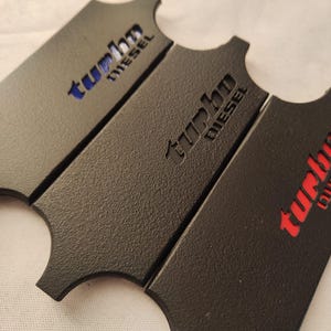 May include: Three black metal plates with a textured finish. Each plate has a unique cutout design and the words "turbo diesel" in different colors: blue, black, and red. The plates are arranged in a slightly overlapping pattern.