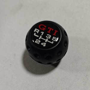 Könnte beinhalten: Schwarzer Schalthebelknauf mit dem Wort "GTI" in Rot und weißem Schaltschema. Der Knauf hat eine geriffelte Oberfläche und ist für ein manuelles Getriebe konzipiert.
