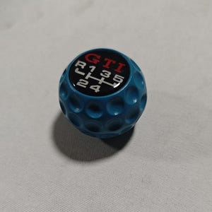 Puede incluir: Un pomo de cambio azul con textura de pelota de golf. La parte superior presenta las letras "GTI" en rojo y un fondo negro con el patrón de cambio de marchas en blanco. Un accesorio para coche.