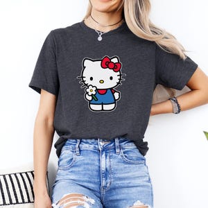 Pu&ograve; includere: Una t-shirt a maniche corte grigio antracite con una stampa grafica del personaggio Hello Kitty. Il personaggio &egrave; bianco con un fiocco rosso, indossa una salopette blu e tiene un piccolo fiore bianco con il centro giallo sul petto.