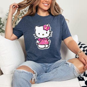 May include: A dark heather blue short-sleeve t-shirt featuring a graphic of a white cartoon cat character with a pink glittery bow and a matching pink glittery dress. The character has black whiskers, a yellow nose, and black outlines.