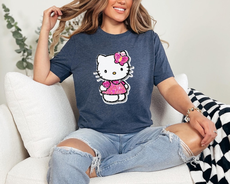 May include: A dark heather blue short-sleeve t-shirt featuring a graphic of a white cartoon cat character with a pink glittery bow and a matching pink glittery dress. The character has black whiskers, a yellow nose, and black outlines.