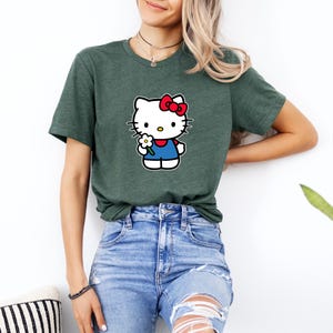 Pu&ograve; includere: Una t-shirt a maniche corte color verde erica scuro con una stampa grafica del personaggio Hello Kitty. Il personaggio &egrave; bianco con un fiocco rosso, indossa un completo blu e tiene un piccolo fiore bianco con il centro giallo sul petto.
