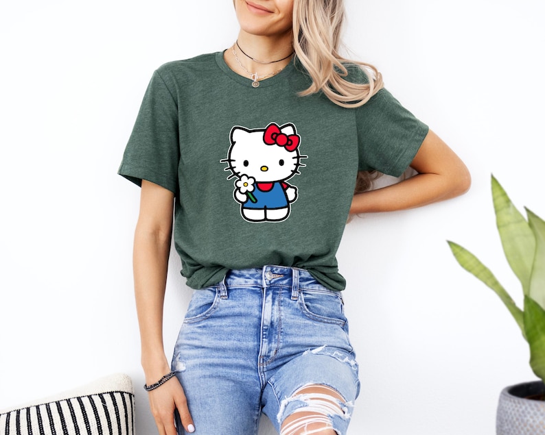 Pu&ograve; includere: Una t-shirt a maniche corte color verde erica scuro con una stampa grafica del personaggio Hello Kitty. Il personaggio &egrave; bianco con un fiocco rosso, indossa un completo blu e tiene un piccolo fiore bianco con il centro giallo sul petto.