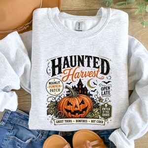 May include: A light gray crewneck sweatshirt featuring a graphic print with the text Haunted Harvest, Moonlit Pumpkin Patch, Open Late, and Enter If You Dare. The design includes a large orange jack-o'-lantern, a dark house, and bats.