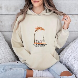 May include: Beige hoodie with a front graphic featuring a tree with orange leaves, characters, and the text "IT'S THE MOST WONDERFUL TIME OF THE YEAR". The hoodie has a drawstring hood and a kangaroo pocket.