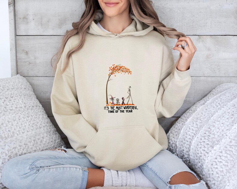 May include: Beige hoodie with a front graphic featuring a tree with orange leaves, characters, and the text "IT'S THE MOST WONDERFUL TIME OF THE YEAR". The hoodie has a drawstring hood and a kangaroo pocket.