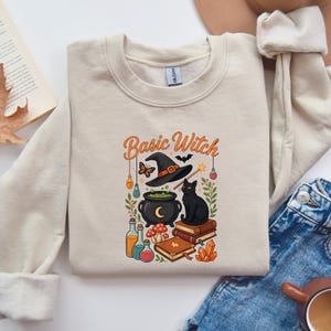 May include: A beige crewneck sweatshirt featuring a graphic print with the text Basic Witch. The design includes a black witch hat, a black cat, a cauldron, books, potion bottles, mushrooms, a butterfly, a bat, and orange crystals.