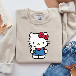 Pu&ograve; includere: Una felpa girocollo color beige con una stampa di Hello Kitty che indossa un fiocco rosso e una salopette blu mentre tiene in mano una margherita bianca. La felpa presenta maniche lunghe con polsini a coste e un'etichetta del marchio Gildan.