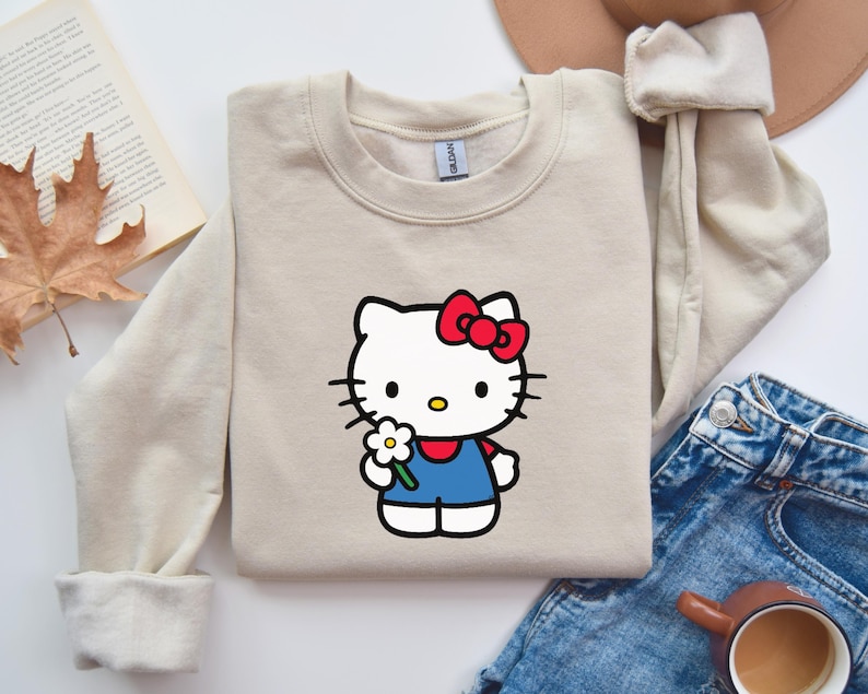 Pu&ograve; includere: Una felpa girocollo color beige con una stampa di Hello Kitty che indossa un fiocco rosso e una salopette blu mentre tiene in mano una margherita bianca. La felpa presenta maniche lunghe con polsini a coste e un'etichetta del marchio Gildan.