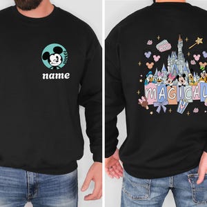 May include: Black sweatshirt featuring Mickey Mouse on the front with the word "name" below. The back of the sweatshirt displays a colorful graphic of a castle, Disney characters, and the word "MAGICAL".